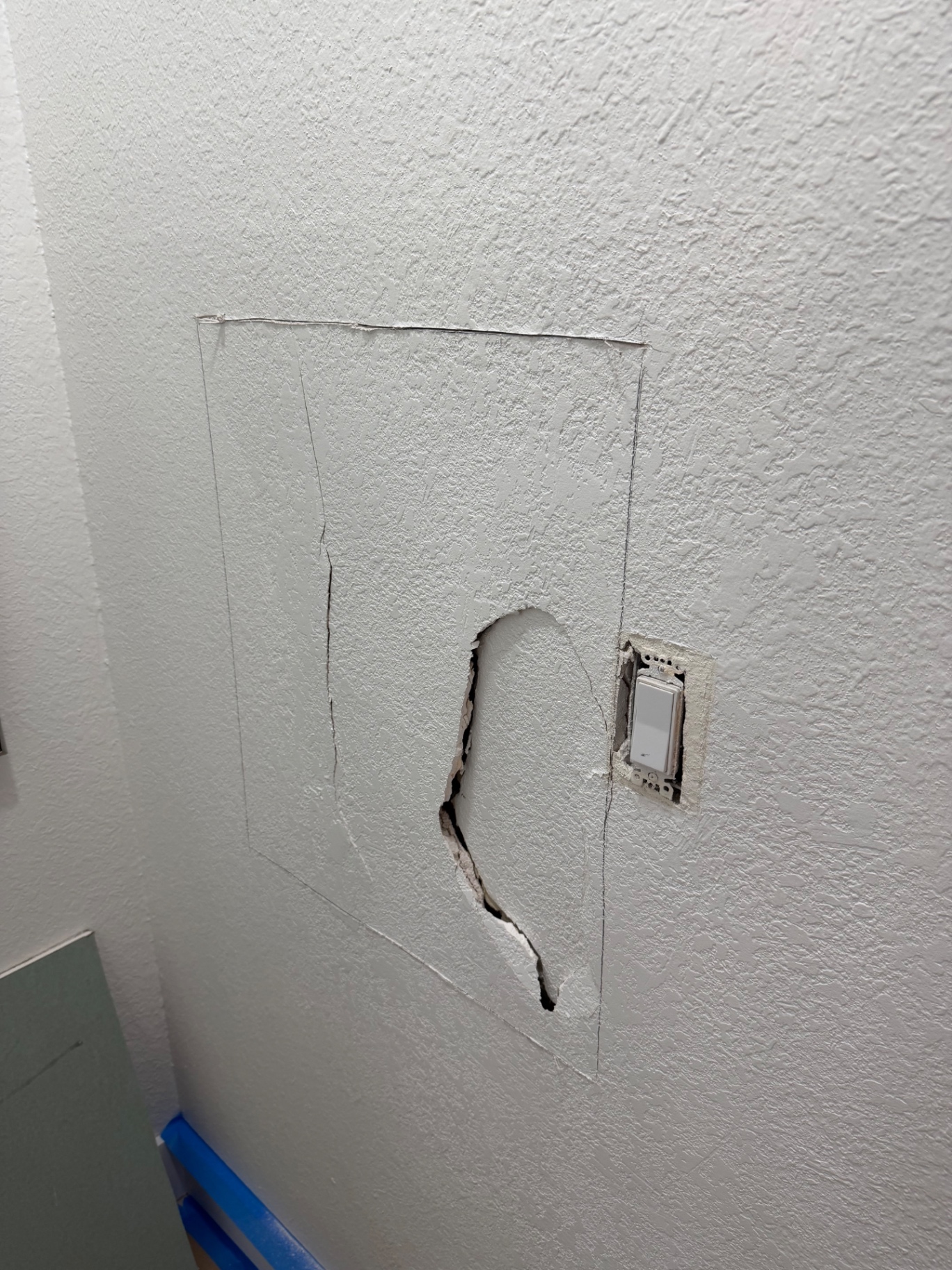 Hole punched through hallway drywall next to light switch, cutout marked for repair